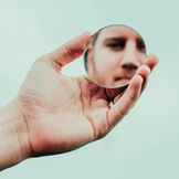 Hand, die einen runden Spiegel hält, in dem das Gesicht einer Person reflektiert wird.